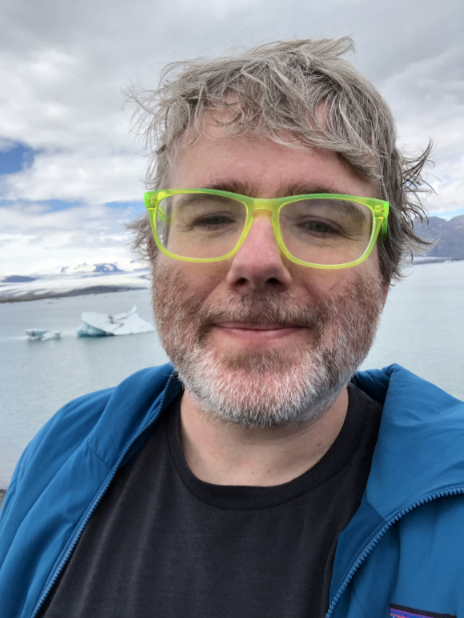 Shaun has grey hair and a grey beard and bright green glasses. In this image he is posing in front of a frozen lake in Iceland.